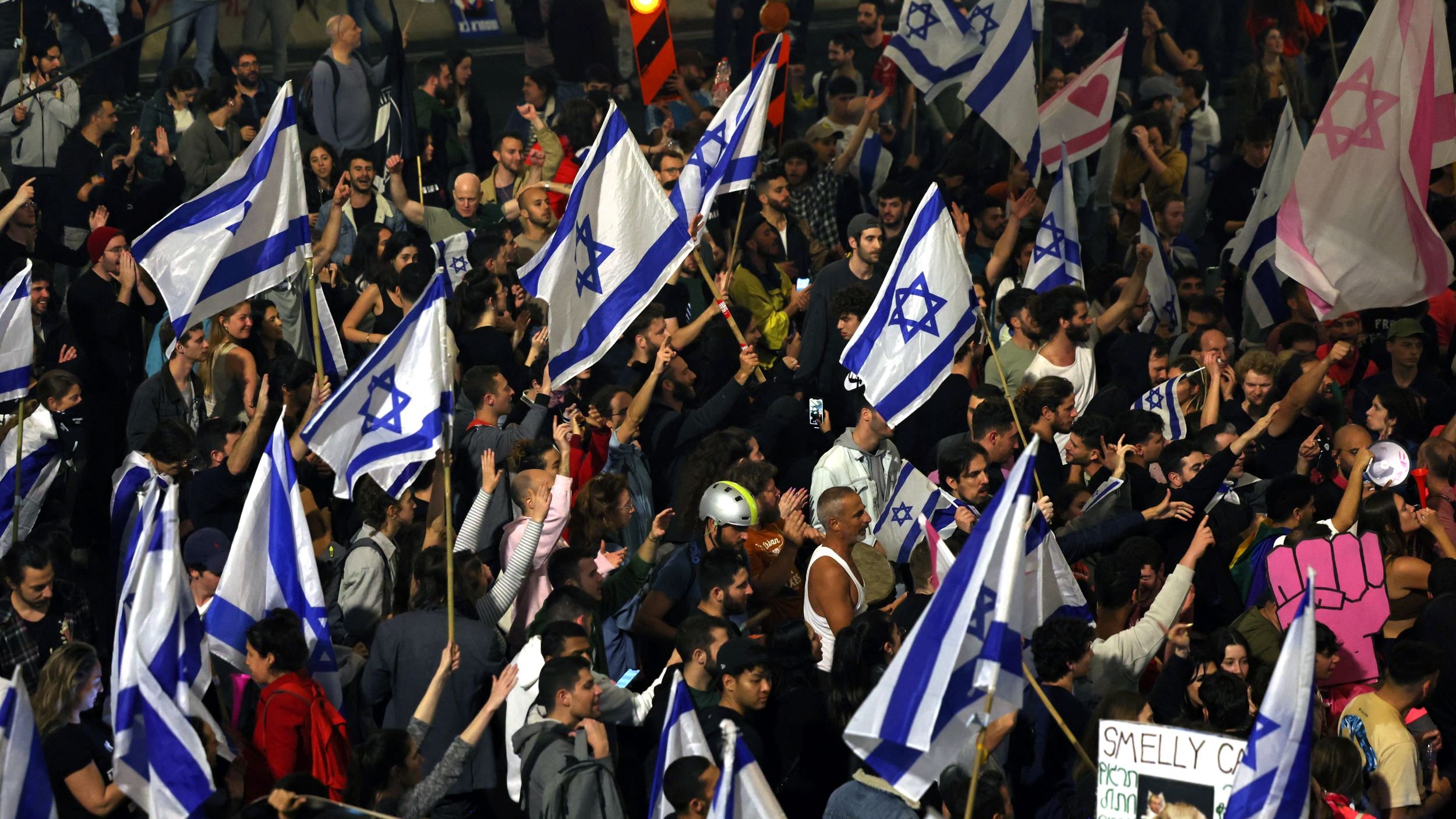 The image depicts a large crowd of people gathered together, many of whom are holding Israeli flags. The atmosphere appears vibrant and energetic, with a mix of participants visible, some raising their hands and signs. Among the signs, one reads "SMELLY C**" in a playful manner. The crowd suggests a protest or demonstration, characterized by a sense of unity and purpose, illuminated by city lights in the background.