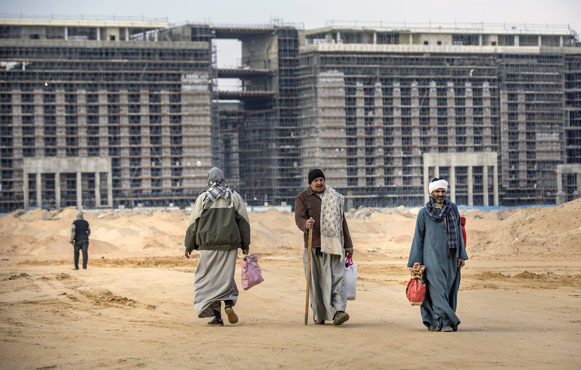 The image depicts a construction site with large, partially completed buildings in the background. In the foreground, four individuals are seen walking on a sandy terrain. They are dressed in traditional attire, including long robes and head coverings. Some are carrying bags, and one person is using a cane. The overall scene reflects a blend of activity and the unfinished nature of the construction, suggesting a developing urban area.