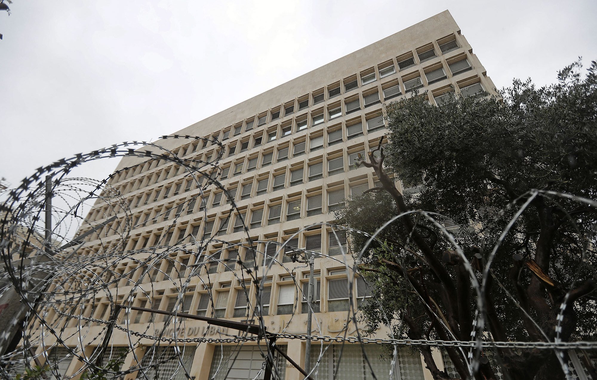 The image shows a large, modern building with a distinct architectural style, featuring multiple windows arranged in a grid pattern. In the foreground, there is a fence topped with barbed wire, indicating a level of security. The scene appears cloudy or overcast, giving a somewhat somber atmosphere. Additionally, there are some trees visible, possibly to the side of the building, adding a touch of greenery to the environment.