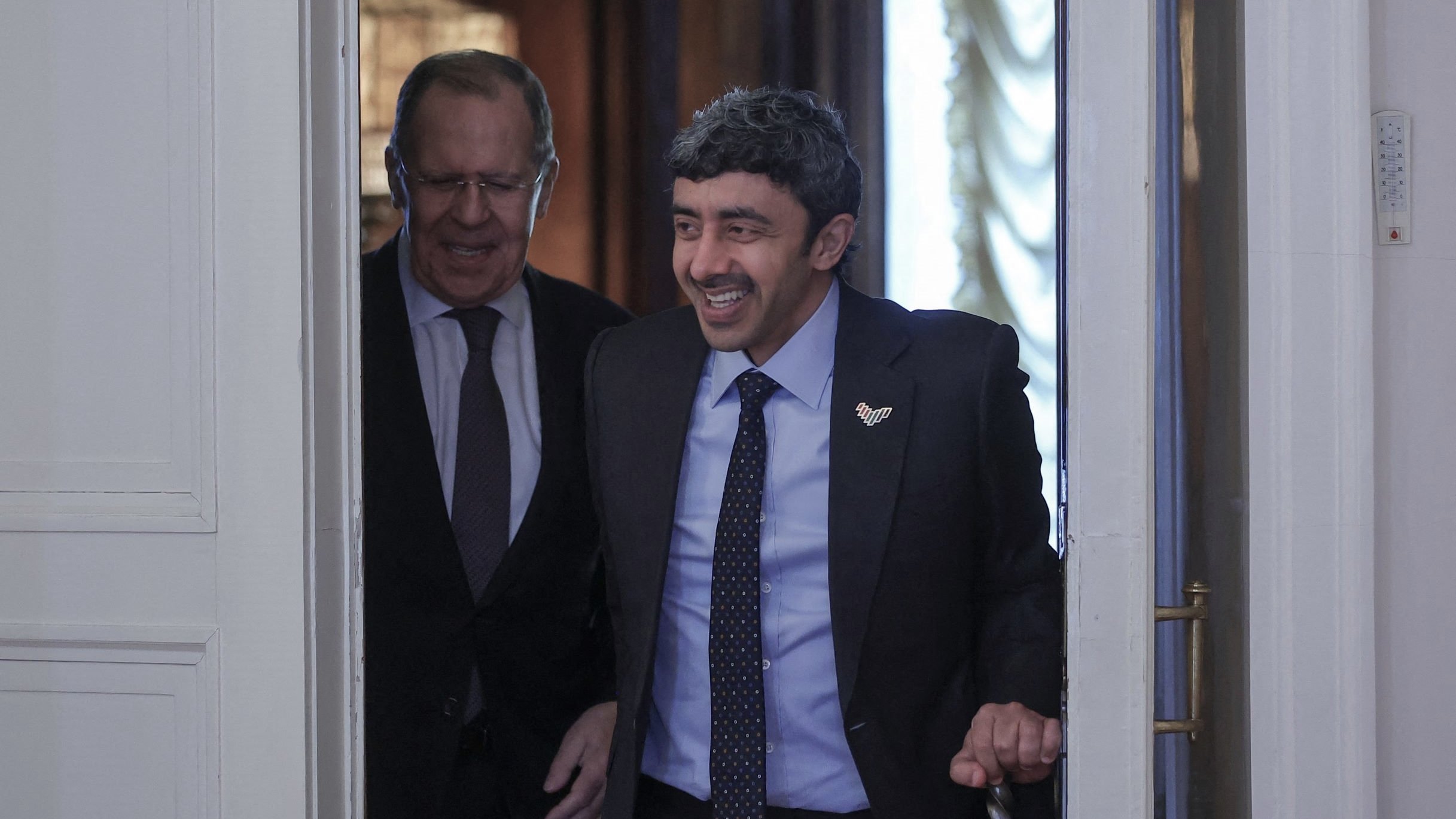 The image shows two men entering a room together. One man is smiling and appears to be in a suit, indicating a formal occasion. The other man, who seems more serious, is walking closely behind. The setting looks elegant, likely a governmental or official location, with decorative elements visible in the background. The overall mood suggests a friendly or cordial interaction between the two individuals.
