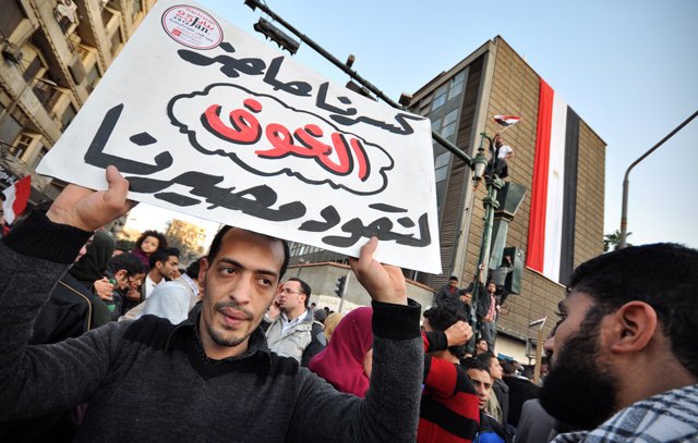 The image depicts a crowd gathered in an urban setting, likely during a protest or demonstration. In the foreground, a man holds a large sign with Arabic text, which translates to "Our demands are not to be ignored." In the background, there is a building displaying the Egyptian flag, suggesting a context related to political or social issues in Egypt. The scene conveys a sense of activism and public expression, with people rallying for their rights or changes in governance.