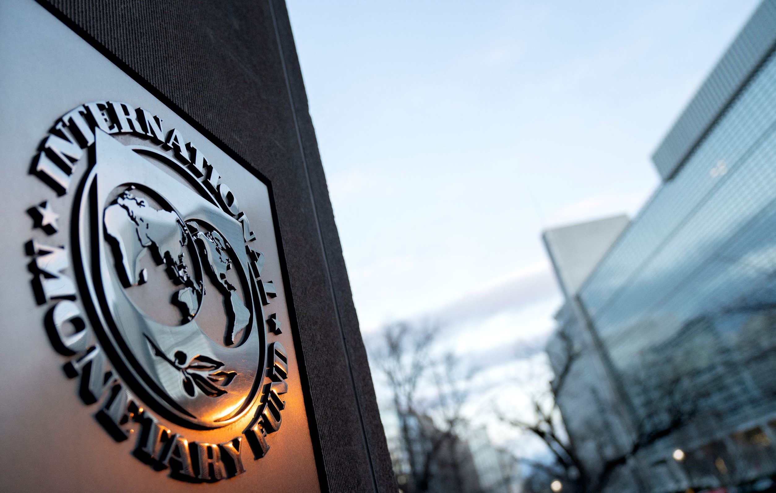 The image features the emblem of the International Monetary Fund (IMF) prominently displayed on a stone surface. The emblem includes a globe surrounded by olive branches, symbolizing peace and cooperation. In the background, there is a modern building with glass windows, reflecting a serene sky with some clouds. The setting appears to be urban and professional, likely outside the IMF headquarters.