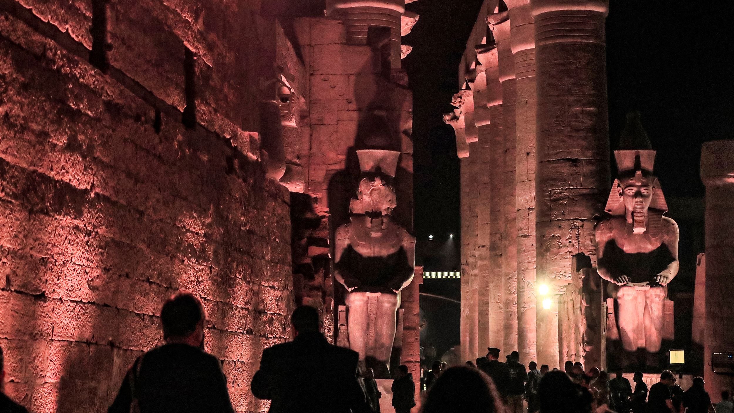 The image depicts a nighttime scene at a historic location, likely a temple or ancient site, characterized by large stone structures. Towering columns line the pathway, illuminated by soft red and amber lighting, which casts dramatic shadows on the walls. Two imposing statues, possibly representing pharaohs or deities, stand prominently at the end of the corridor. Visitors can be seen walking through the area, taking in the majestic architecture and ambiance of this ancient site. The atmosphere conveys a sense of history and reverence.
