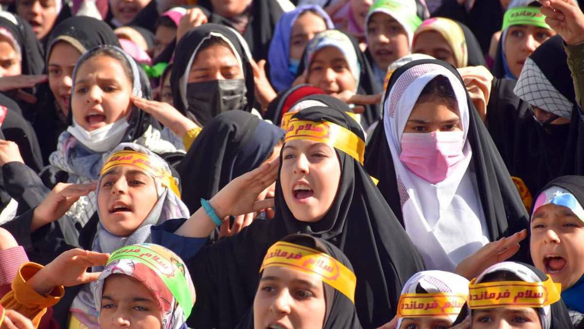 The image shows a large group of girls and young women gathered together, likely participating in an event or demonstration. They are wearing colorful headscarves and black clothing, along with yellow headbands that feature Arabic text. The girls appear to be actively engaged, singing or chanting, and expressing enthusiasm. Their expressions convey a sense of unity and purpose as they participate in this collective activity.