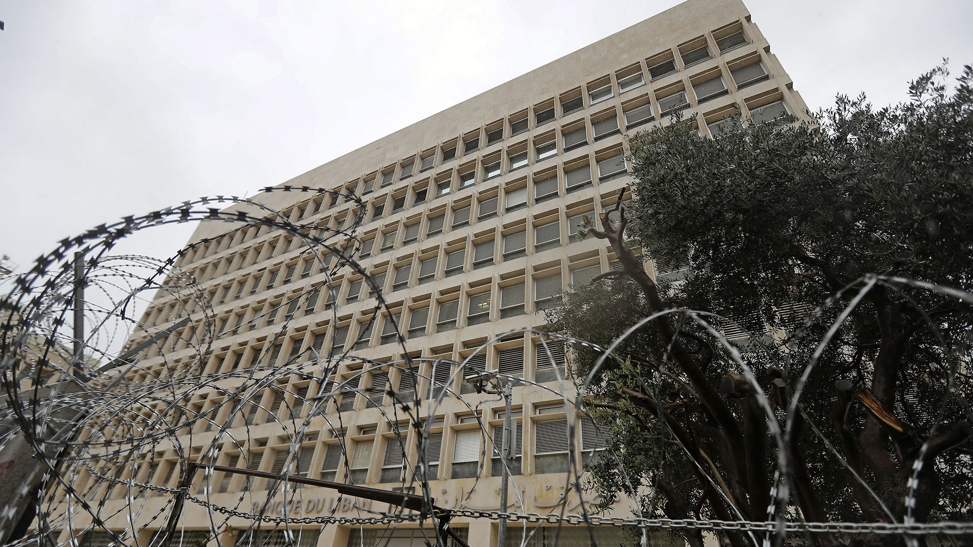The image shows a large, modern building with a distinct architectural style, featuring multiple windows arranged in a grid pattern. In the foreground, there is a fence topped with barbed wire, indicating a level of security. The scene appears cloudy or overcast, giving a somewhat somber atmosphere. Additionally, there are some trees visible, possibly to the side of the building, adding a touch of greenery to the environment.