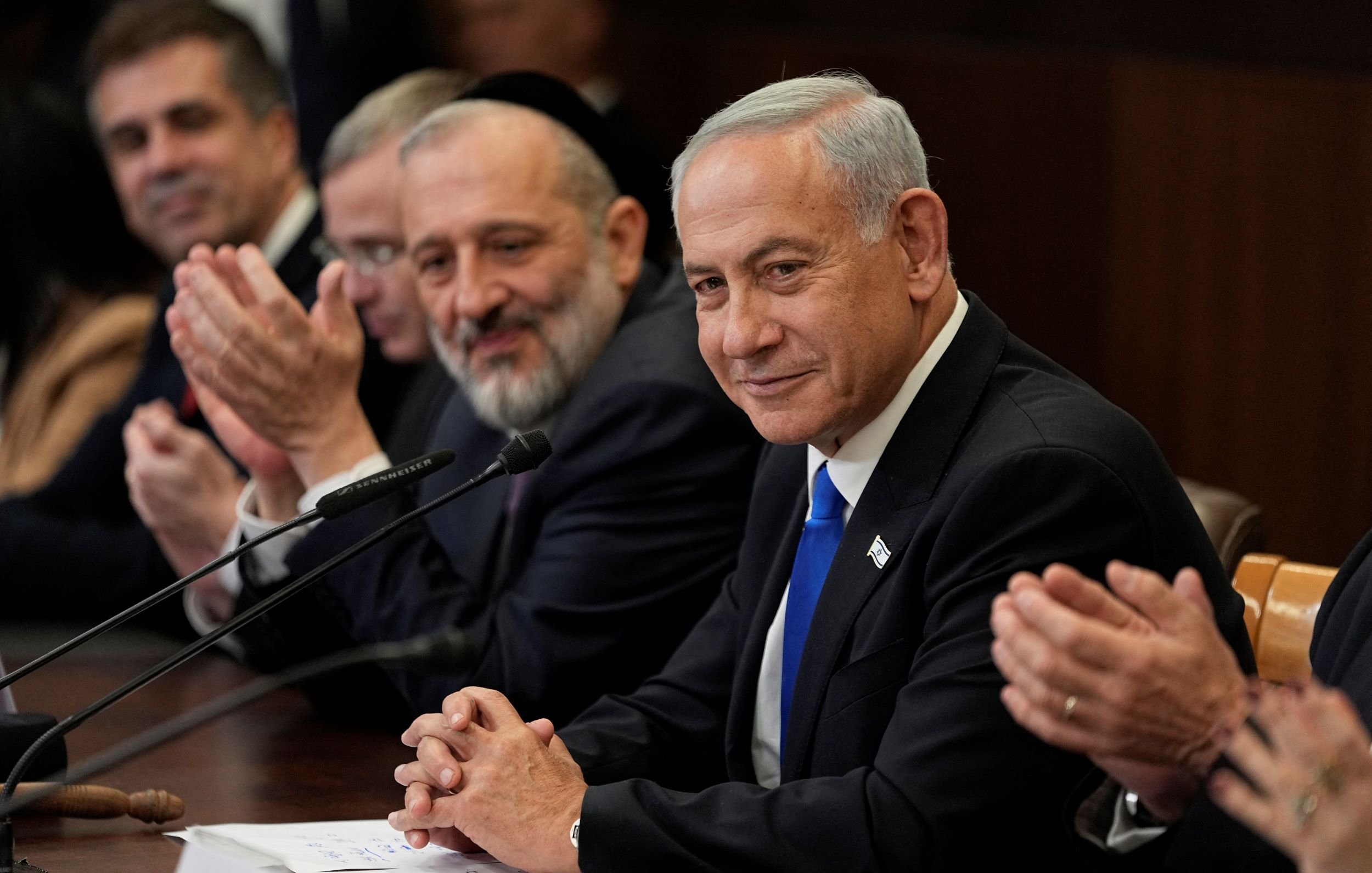 The image depicts a group of men seated around a table during a formal meeting. In the foreground, a man in a suit with a blue tie appears to be the focal point, wearing a slight smile. He is flanked by other men who are also dressed in formal attire. One man, likely a religious figure, is seen smiling broadly. The setting appears to be an official governmental or diplomatic environment, indicated by the wooden table and the serious demeanor of those present. There are papers and a microphone in front of them, suggesting they are engaged in a discussion or announcement.