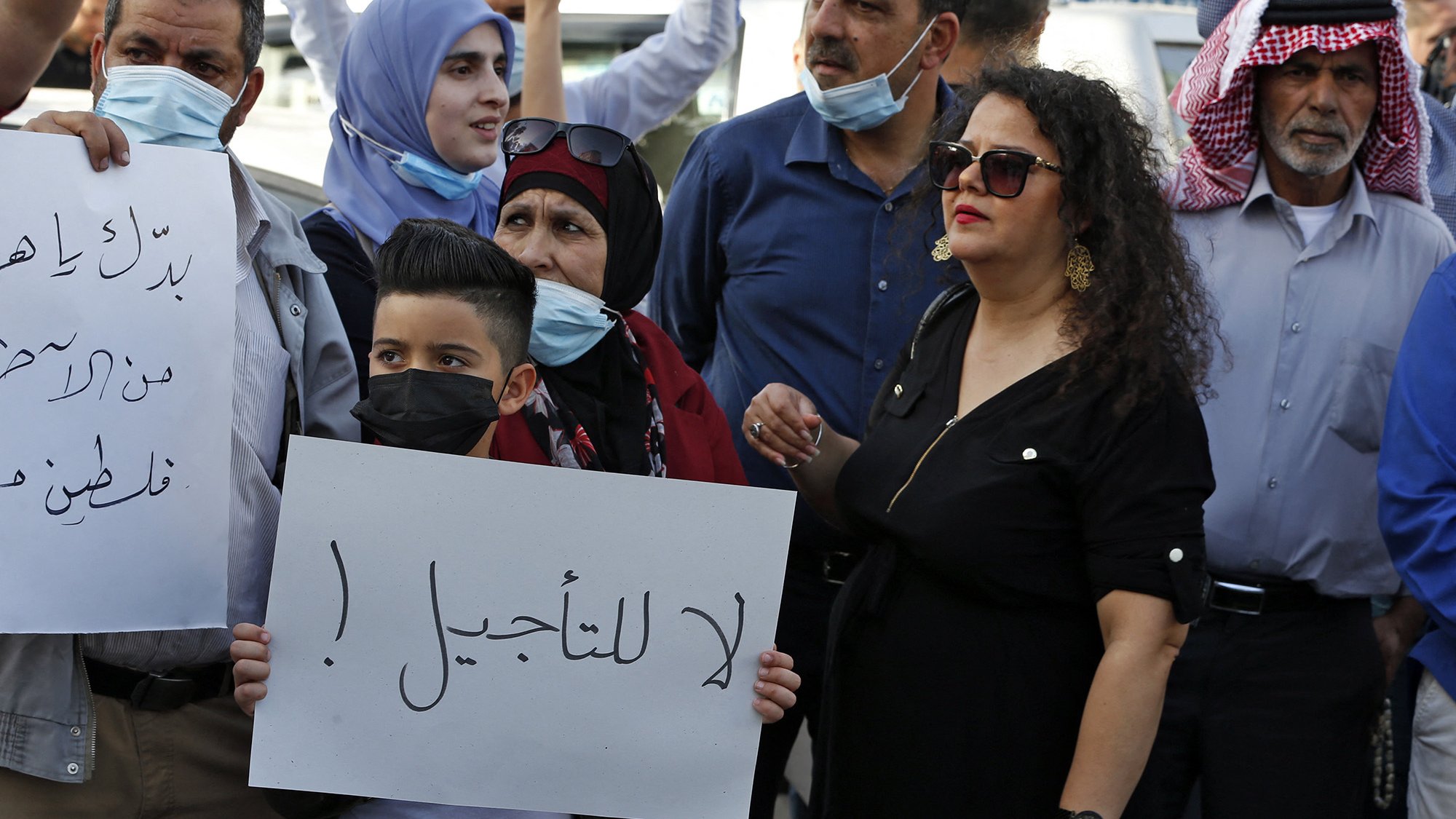 The image depicts a group of people participating in a protest. Some individuals are wearing masks, and they are holding signs, including one that prominently features the Arabic phrase "لا للتأجيل" (translated as "No to delay"). The crowd appears to include men, women, and children, with a mix of facial expressions that suggest determination and solidarity. The background shows a public space, likely in an urban setting.