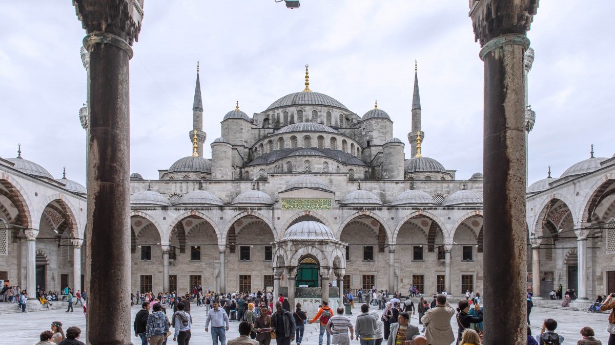 The image features a grand architectural view of the Sultan Ahmed Mosque, commonly known as the Blue Mosque, in Istanbul, Turkey. In the foreground, you can see a bustling courtyard filled with visitors and tourists. The mosque's impressive structure is visible in the background, characterized by its large dome and multiple tall minarets reaching upward. The scene is framed by arched columns, adding to the architectural beauty. The sky appears overcast, creating a soft light that enhances the details of the mosque's intricate design.