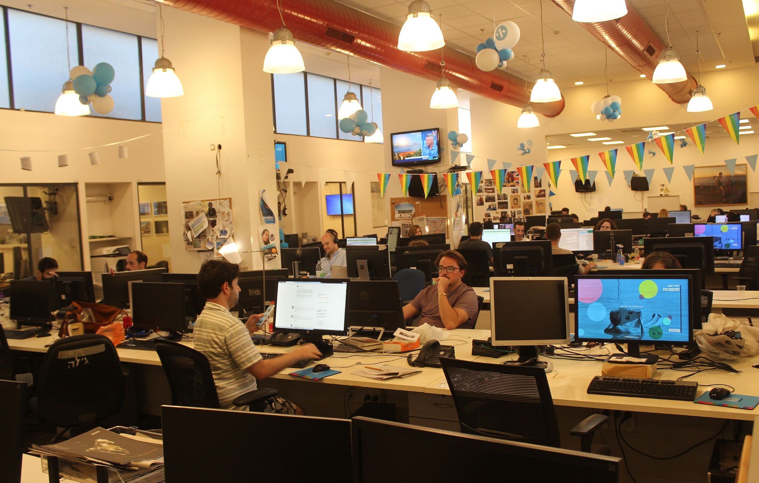 The image shows an open office space with several workstations. There are multiple desks equipped with computers, where people are engaged in various tasks. The decor includes colorful balloons and banners hanging from the ceiling, suggesting a festive atmosphere. Large windows allow for ample natural light, and the overall ambiance seems lively and collaborative, with employees working both individually and in small groups.
