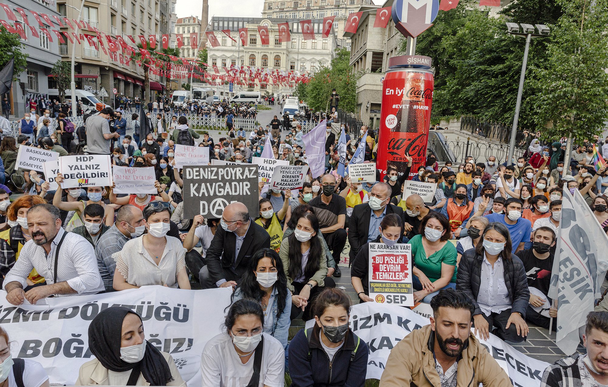The image depicts a large gathering of people participating in a protest. Many of the individuals are wearing masks and holding various signs and banners with slogans, indicating their support for a cause. The scene appears to be in an urban setting, likely a city center, with flags in the background. The participants seem to be sitting on the ground, emphasizing their determination and solidarity for the issue they are advocating. The atmosphere looks charged with activism and communal spirit.