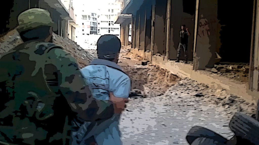 The image depicts a war-torn urban environment. A soldier in military attire is seen guiding or restraining a person wearing a light-colored shirt. The setting suggests destruction, with debris scattered on the ground and the remnants of buildings in disrepair. In the background, another individual is partially visible, possibly further down the street. The overall atmosphere conveys a sense of tension and conflict.
