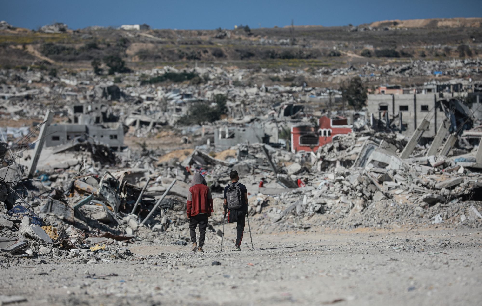 Two people walk through a vast landscape of destruction and rubble, with collapsed buildings around them.