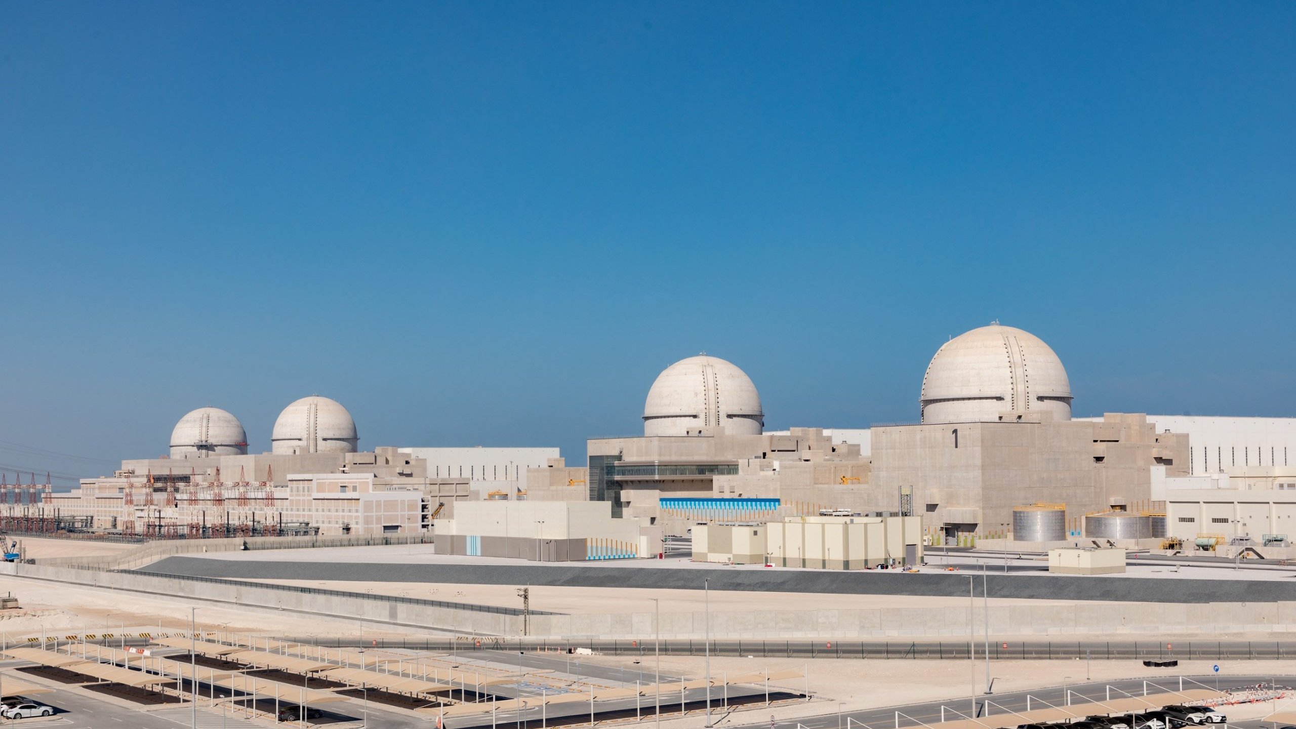 The image shows a large industrial facility, likely a nuclear power plant, with several prominent dome-shaped structures. The plant features a modern design with various buildings and infrastructure surrounding it, set against a clear blue sky. The setting appears to be an arid or semi-arid environment, with minimal vegetation. There are parking areas and some infrastructure elements visible in the foreground, indicating it is a well-developed site.