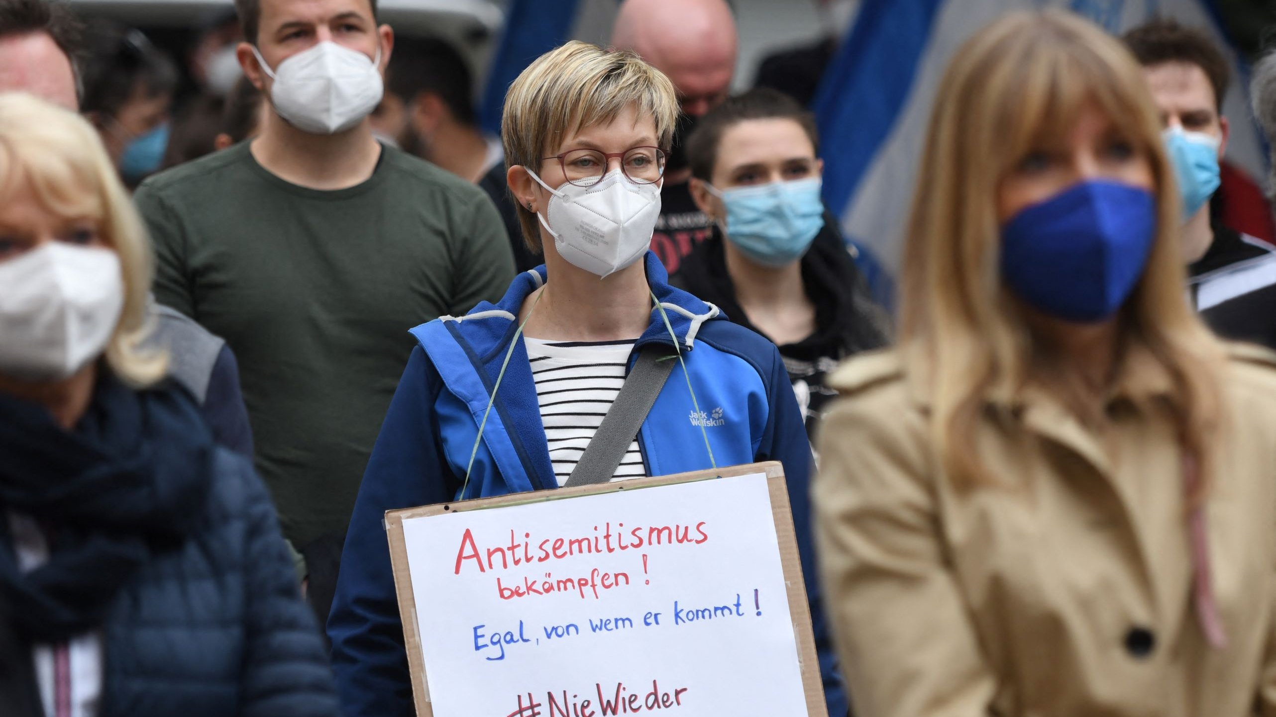 The image shows a group of people gathered at an outdoor event or protest. In the foreground, a woman wearing a blue jacket and a mask holds a sign that reads "Antisemitismus bekämpfen!" (which translates to "Fight antisemitism!") and "Egal, von wem er kommt!" (meaning "No matter who it comes from!"). The hashtag "#NieWieder" is also visible on the sign. The crowd appears to be composed of individuals also wearing masks, reflecting a collective stance on the issue at hand. The setting seems to be a public demonstration focused on combating antisemitism.