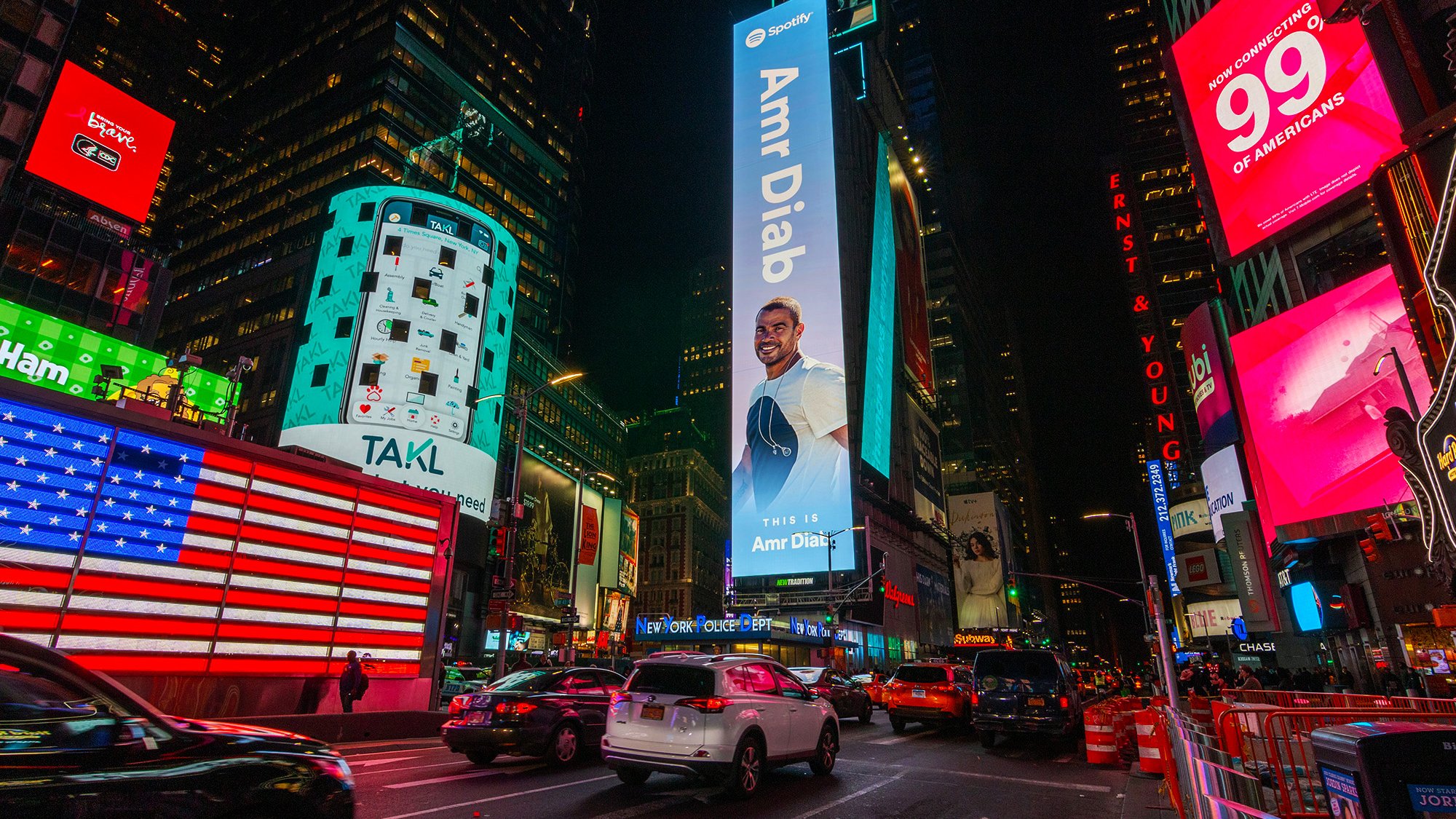 The image depicts a vibrant scene in Times Square, New York City, at night. The area is illuminated by numerous bright advertisements and digital billboards. Prominently featured is a large advertisement for Amr Diab, an acclaimed musician, which invites viewers to listen to his music. There is an American flag displayed on one of the buildings, and several vehicles are seen navigating the busy street. The atmosphere is lively, characteristic of this iconic urban location.