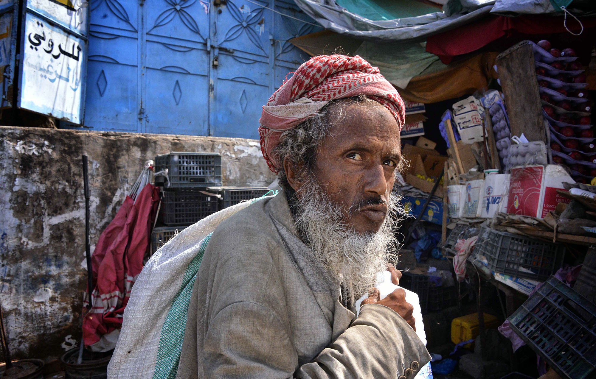 The image depicts an elderly man with a prominent beard and traditional attire, wearing a distinctive head wrap. He appears to be in a market setting surrounded by various goods and a colorful backdrop. His expression seems contemplative, and he is holding a container, possibly of milk or another liquid. The scene conveys a sense of everyday life in a bustling environment.