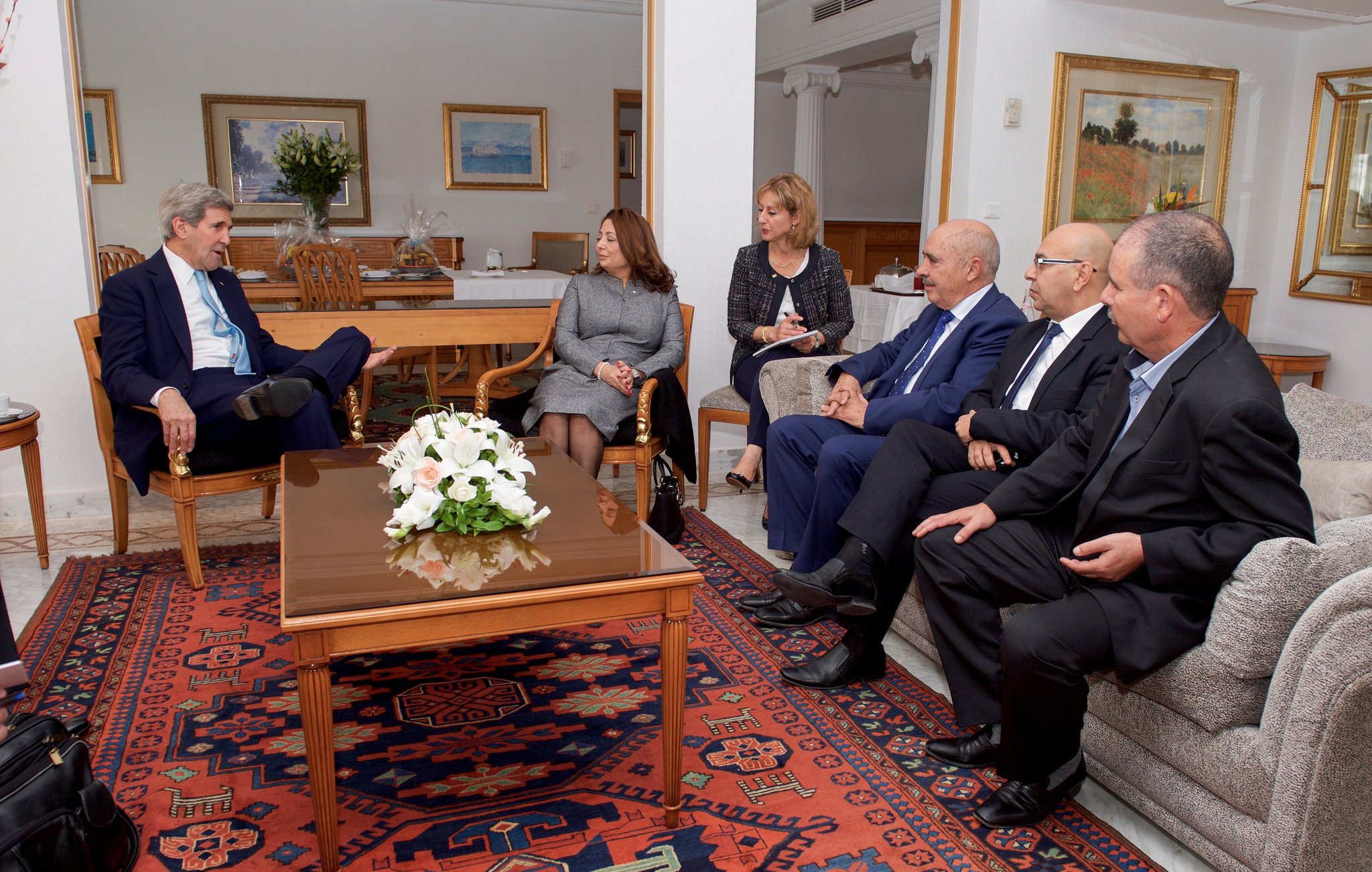 The image depicts a formal meeting taking place in a well-lit, elegantly furnished room. A group of professionals is seated around a coffee table, engaged in discussion. On one side, a person in a suit appears to be leading the conversation, while others listen attentively. The room features tasteful decor, including paintings on the walls and a floral arrangement on the table, creating a calm and sophisticated atmosphere. The group consists of both men and women, indicating a diverse representation.