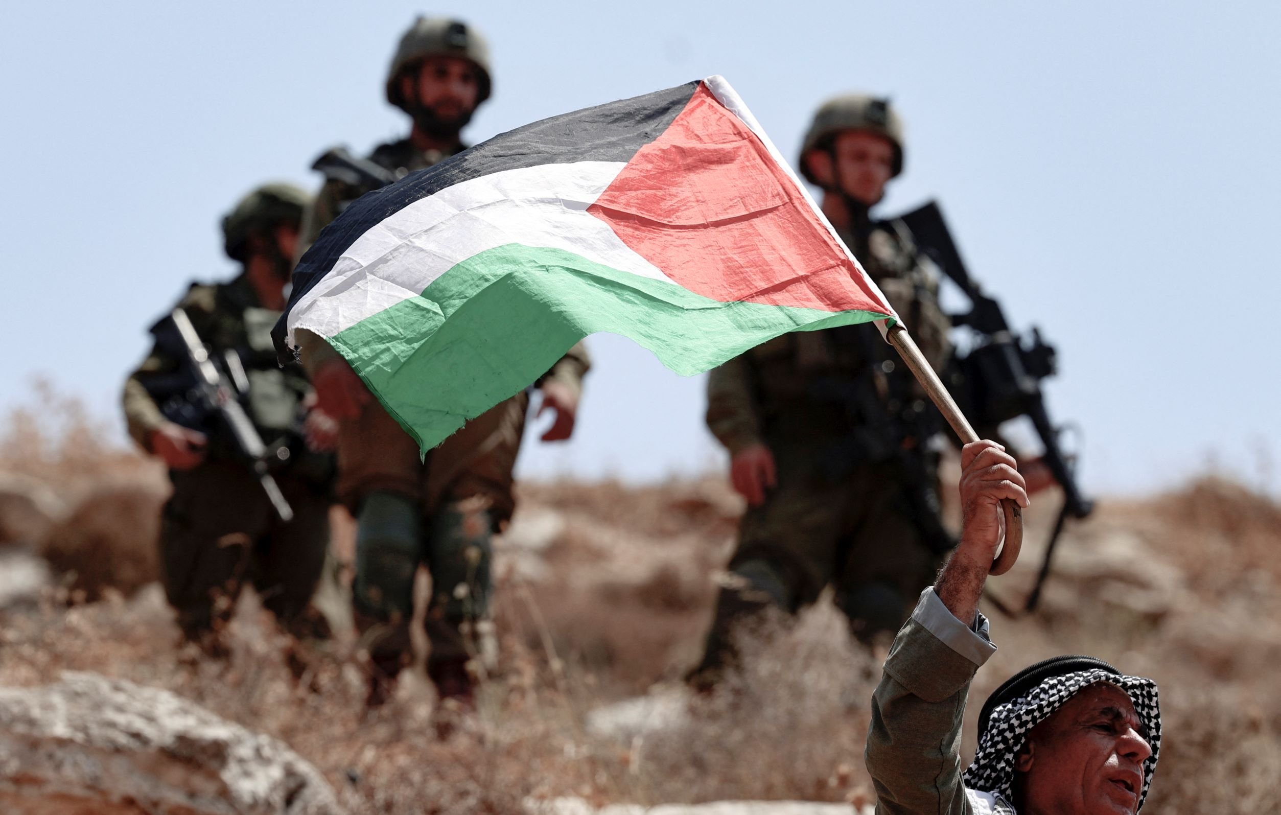 The image shows a man holding a Palestinian flag aloft, standing in a rocky landscape. He appears to be expressing a strong patriotic sentiment. In the background, a group of soldiers is visible, dressed in military gear, suggesting a tense or conflict-related scene. The setting looks arid, typically associated with certain regions in Palestine. The overall atmosphere conveys a sense of tension and resistance.