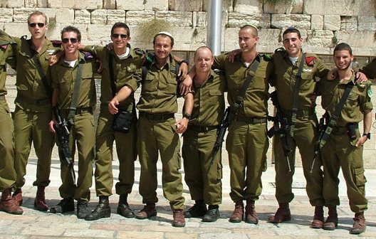 The image features a group of eleven soldiers standing closely together, wearing olive green military uniforms and boots. They appear to be in a celebratory mood, with smiles and casual poses. The background suggests a stone wall, possibly the Western Wall in Jerusalem, indicating a significant location. The soldiers carry some equipment, hinting at their military roles. Overall, the image conveys camaraderie and a sense of accomplishment among the group.