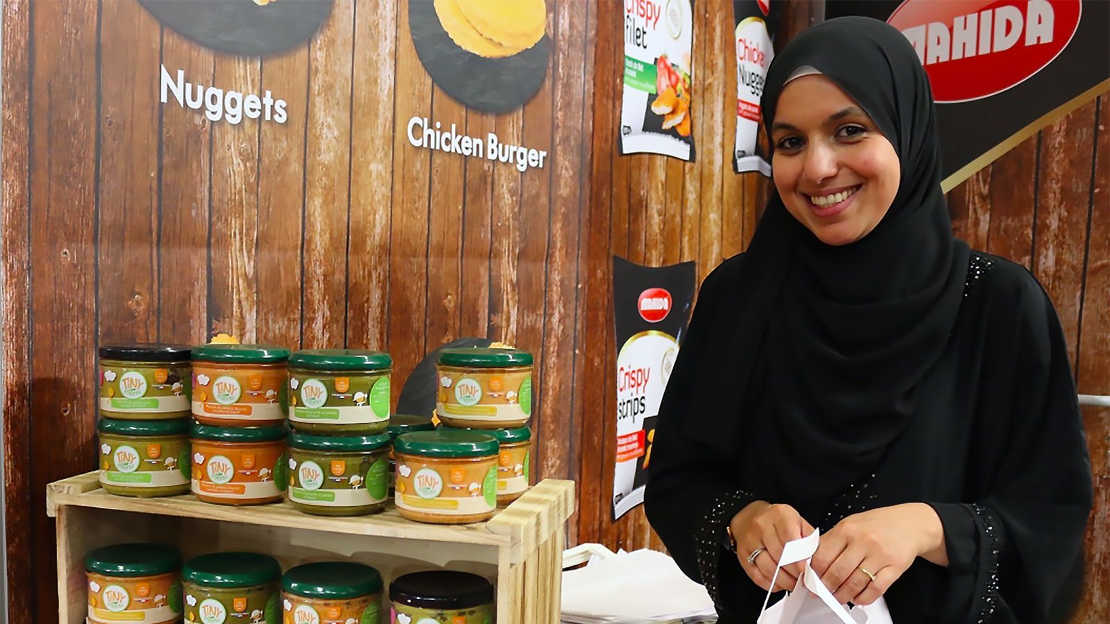 The image features a woman wearing a black hijab, smiling while standing behind a display of jars on a wooden table. The jars are arranged in stacks, showcasing various products, including flavors such as nuggets and chicken burger. The background has a wooden texture, and there are promotional posters or signs visible. The overall setting suggests a market or food exhibition atmosphere.