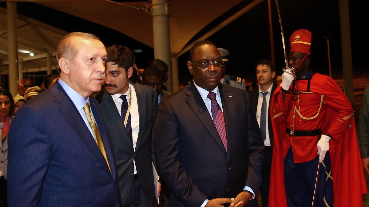 The image shows two prominent figures walking together in a formal setting. One is wearing a dark suit with a light blue striped tie, and the other is dressed in a dark suit with a maroon tie. They appear to be engaged in conversation or deep in thought. In the background, there is a ceremonial guard dressed in a bright red uniform, standing at attention, adding a sense of formality to the scene. The environment looks like an official venue, possibly an airport or a conference center, illuminated by soft lighting.