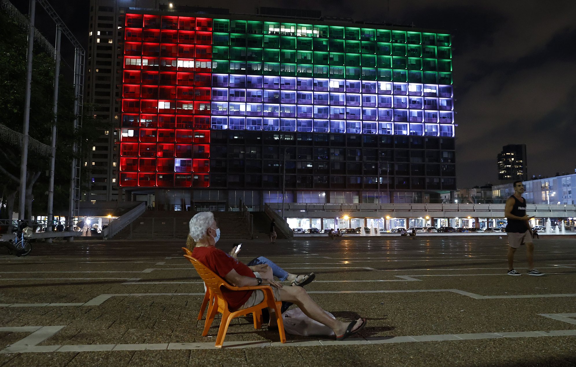 The image shows a large building illuminated with colorful lights at night, displaying the colors of a flag: red, green, and white. In the foreground, an elderly man is sitting in a plastic chair, looking at his phone. The scene conveys a sense of tranquility, with the illuminated building serving as a striking backdrop in a public square. In the background, other people can be seen walking or engaging in activities. The atmosphere appears calm and reflective.