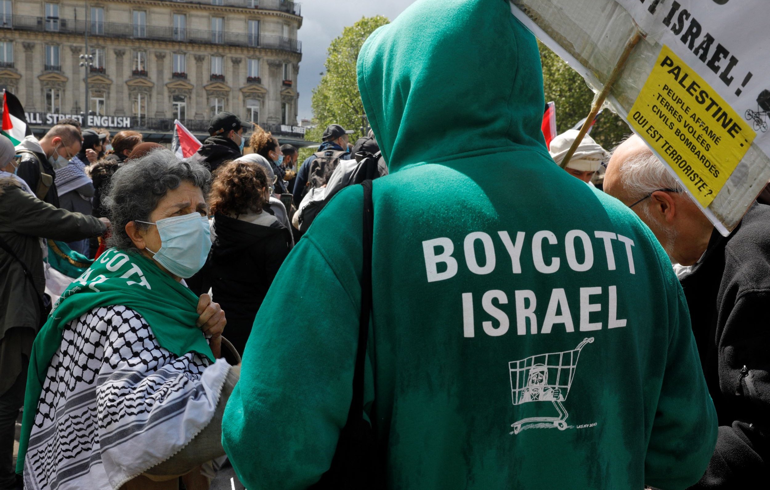 The image depicts a gathering or protest where individuals are expressing their political views. In the foreground, a person wearing a green hoodie that reads "BOYCOTT ISRAEL" is facing another individual. The latter is wearing a face mask and has a traditional black-and-white patterned scarf. The setting appears to be in a public space, with banners and flags present in the background, indicating a demonstration related to Palestinian rights or opposition to certain policies regarding Israel.