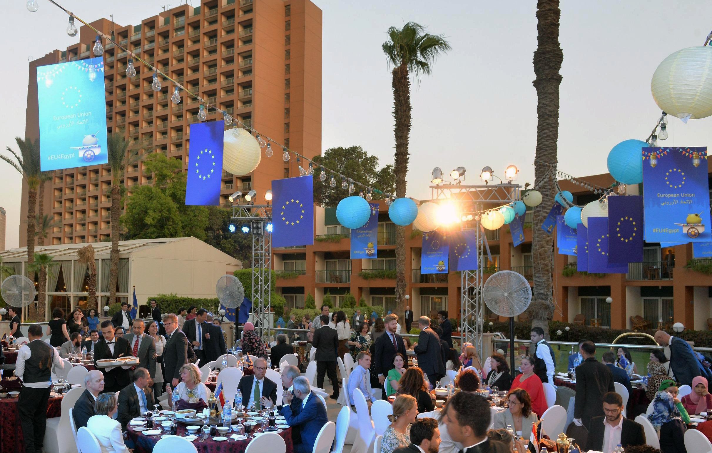 The image depicts an outdoor event, likely a formal gathering or dinner party set up in a picturesque location. Tables are elegantly arranged with white tablecloths and chairs, and guests are dressed in formal attire, engaging in conversation. The ambiance is enhanced by decorative blue flags, likely representing the European Union, and hanging lanterns that illuminate the scene. Palm trees are visible in the background, adding a tropical feel, while a hotel or building looms in the background. The atmosphere appears lively and festive, suggesting a celebration or special occasion.
