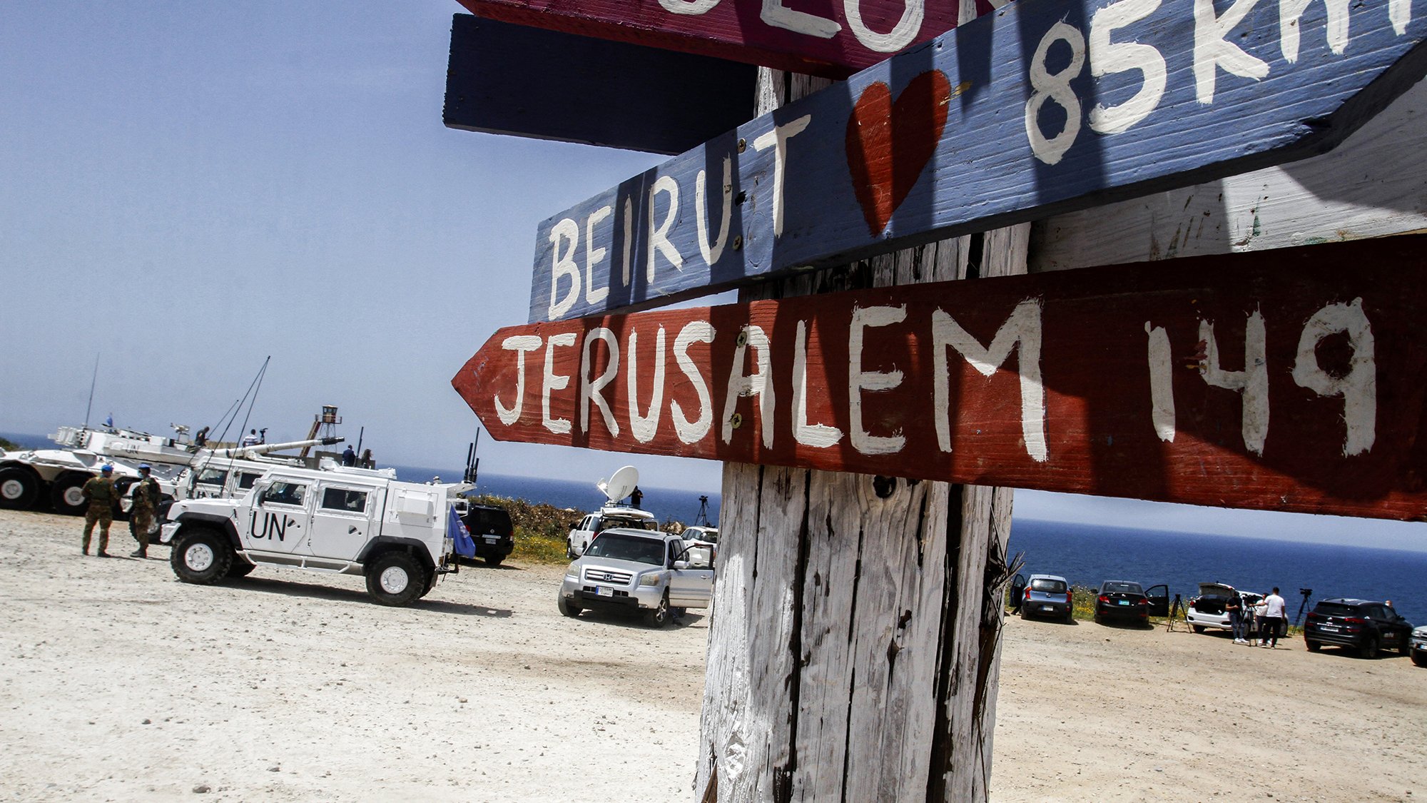 The image depicts a wooden signpost with several directional arrows pointing towards different locations. The names of the places, including "Beirut" and "Jerusalem," are painted in bold, colorful letters. The distances to these locations are also indicated in kilometers. In the background, there are vehicles, including a UN truck, parked on a sandy area near the coastline, with a view of the sea. The overall setting appears to be a scenic lookout or border area.