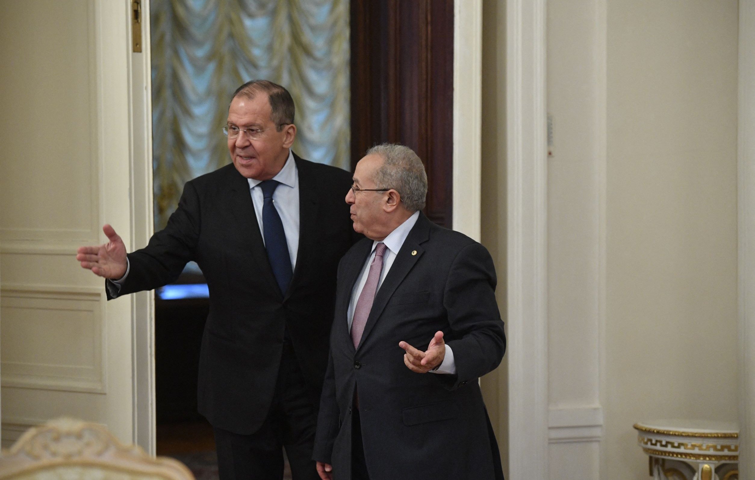 The image shows two men in a formal setting, likely inside a government building. One man is gesturing with his hand as he walks, indicating he is speaking or inviting the other man to follow. The second man appears to be listening attentively. Both are dressed in formal attire, suggesting a diplomatic or official meeting. The room has a sophisticated decor, with elements such as elegant walls and furniture visible in the background.