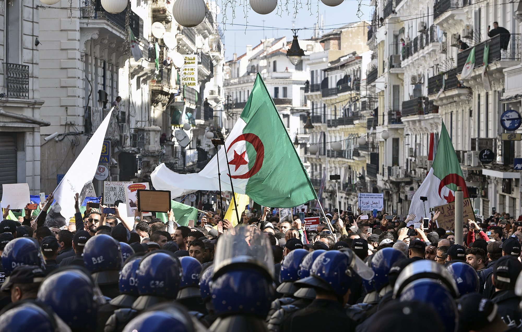 The image depicts a large protest scene in a street lined with buildings. Many people are gathered, holding flags, including Algerian flags, and signs. The crowd is diverse and appears to be demonstrating for a cause. In the foreground, a line of riot police in helmets is visible, standing in front of the protesters, indicating a tense atmosphere. The street is adorned with lights and decorations, suggesting it might be a significant location in the city. Overall, the image captures a moment of civic engagement and protest.