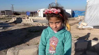 The image depicts a young girl with striking blue eyes and dark hair, wearing a bright turquoise sweatshirt adorned with a character design on the front. She has a decorative pink headband with flowers, which adds a playful touch to her appearance. The background shows a makeshift camp with tents and some structures, indicating a setting that may be temporary or in a challenging environment. The girl is looking directly at the camera with a slight smile, suggesting a sense of resilience and charm amidst her surroundings.