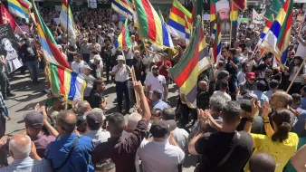 The image depicts a large crowd gathered in a public space, participating in a demonstration or protest. Many people are holding brightly colored flags that feature a mix of horizontal and vertical stripes. The atmosphere appears energetic and spirited, with participants actively engaging with one another. Some individuals are raising their hands and clapping, suggesting a celebratory or passionate mood. In the background, various signs and posters can be seen, indicating that the event is likely focused on a specific cause or issue.