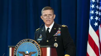 The image shows a man in a military uniform standing at a podium. The podium has the emblem of the Department of Defense prominently displayed. The man is speaking, possibly at a formal event or press conference. Behind him, there is a blue backdrop, and an American flag is visible to the side. The setting appears to be official and ceremonial.