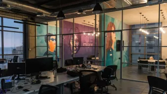 The image shows a modern office space with large glass windows, providing a view of the outside. Inside, there are several workstations with computers and ergonomic chairs. The walls are decorated with vibrant murals featuring stylized portraits of individuals, adding a creative touch to the environment. The overall ambiance is open and contemporary, with industrial elements like exposed ducts and warm lighting fixtures. The space appears to be designed for collaboration and innovation.