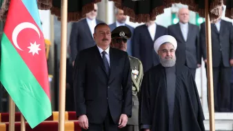 The image depicts a formal meeting between two leaders, presumably from Azerbaijan and Iran, standing side by side. In the foreground, one leader is dressed in a suit, while the other is wearing a dark robe and headpiece. Behind them, a group of individuals can be seen, and a large flag of Azerbaijan is displayed prominently on the left. The setting appears to be an official location, likely a government building, with stairs and a red carpet visible.