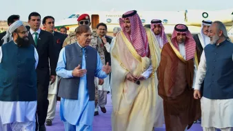 The image depicts a group of men walking together, likely during a formal event or diplomatic visit. They are dressed in traditional attire, with some wearing long robes and others in suits. The setting appears to be an airport or other official venue, indicated by the presence of a private jet in the background. The men seem engaged in conversation, and the atmosphere suggests camaraderie or collaboration. The purple carpet hints at a ceremonial welcome.
