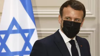 The image shows a man in a dark suit wearing a black face mask. He appears to be looking slightly to the side, with a serious expression. In the background, there is a flag featuring a blue Star of David on a white field, which likely signifies that the setting is related to Israel. The overall atmosphere suggests a formal or diplomatic context.