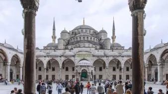 The image features a grand architectural view of the Sultan Ahmed Mosque, commonly known as the Blue Mosque, in Istanbul, Turkey. In the foreground, you can see a bustling courtyard filled with visitors and tourists. The mosque's impressive structure is visible in the background, characterized by its large dome and multiple tall minarets reaching upward. The scene is framed by arched columns, adding to the architectural beauty. The sky appears overcast, creating a soft light that enhances the details of the mosque's intricate design.