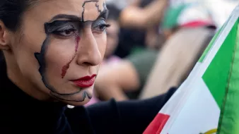 The image features a woman with striking face makeup that outlines the map of a country, highlighting it with dark lines. She has a tear streaking down her cheek, adding an emotional touch to her expression. The woman holds a flag that includes green, white, and red colors, which likely represents her national identity. The setting indicates a public gathering or protest, as there are people in the background, contributing to a vibrant atmosphere.