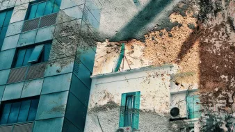 The image features a juxtaposition of two buildings. On the left, there is a modern structure with large blue-tinted windows and metal panels. On the right, an older building displays a weathered wall with patches of peeling paint and exposed textures. A couple of small windows with turquoise shutters add color to the aged facade. Additionally, air conditioning units are mounted on the wall, contrasting the contemporary look of the building on the left with the rustic charm of the one on the right. The overall color palette is a mix of cool blues and warm earthy tones.