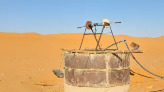 The image shows an old water well situated in a vast desert landscape. The well is made from a large, cylindrical structure with a rusty metal exterior and a concrete base. At the top, there is a simple wooden frame holding a pulley and a couple of wheels, likely used for drawing water. Surrounding the well are sand dunes, displaying a warm golden color under a clear blue sky, emphasizing the arid environment.