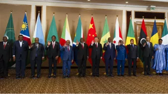 The image features a group of politicians standing together in a formal setting. They are arranged in a line, with each person smiling and waving. Behind them, there are several national flags, including those of various countries such as China and several African nations, indicating an international or diplomatic event. The individuals appear to represent different countries or organizations, reflecting a multicultural gathering. The setting has an elegant interior, suggesting a significant and official occasion.