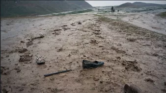 The image depicts a muddy landscape, likely after rainfall. The ground is uneven and dotted with puddles and dirt. Scattered across the terrain are several shoes, including a black dress shoe and what appears to be a wooden shoe or clog. There is also a long, thin object that resembles a cane or stick. The scene conveys a sense of abandonment and desolation, with lush green hills in the background under a gray sky.