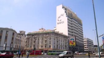 The image shows a cityscape featuring a tall building with the letters "BMCI" prominently displayed on it. The architecture of the surrounding buildings is a mix of modern and traditional styles. The sky is clear and blue, indicating a sunny day. In the foreground, there are vehicles on the road, including a red car and a few buses, as well as a billboard advertising various products. The scene conveys a bustling urban environment.