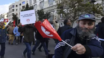 The image depicts a protest scene, likely in Tunisia. A crowd of people is marching down a street. Some participants are holding signs and flags. One prominent sign reads "ALL UNITED AGAINST DICTATORSHIP!" indicating the protest's focus on political issues. Among the crowd, a man stands out, waving a Tunisian flag and wearing a baseball cap and a jacket. The overall atmosphere suggests a movement against authoritarian rule, with participants expressing their collective dissent.