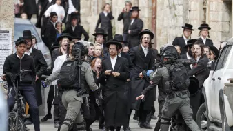 The image depicts a tense confrontation between a group of individuals dressed in traditional Orthodox Jewish attire and armed security personnel. The group appears to be expressing strong emotions, possibly protest or unrest, while the security officers, equipped with tactical gear, seem to be engaging with them. The setting appears to be an urban area, with a mixture of traditional and modern elements visible in the background, including vehicles and stone buildings. The scene captures a moment of conflict or significant social interaction.