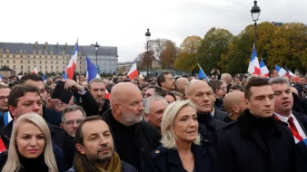 The image depicts a crowd of people gathered in an outdoor setting, likely at a political event or rally. Many individuals are holding French flags, and the expressions on their faces suggest a mix of determination and focus. Prominent figures can be seen at the forefront, wearing formal attire. The background features historic architecture and trees, indicating an urban environment. The atmosphere appears charged, reflecting the significance of the gathering.