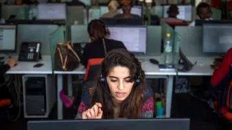 The image depicts a call center environment. In the foreground, a young woman is wearing a headset, concentrating intently on her work at a computer. She appears to be engaged in a conversation, with one hand raised, possibly indicating she is speaking or asking a question. In the background, several other people are working at similar desk setups, each using computers and headsets, which creates a bustling atmosphere. The workspace is well-lit and modern, with a mix of personal items placed at some desks.
