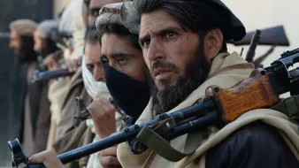 The image features a group of individuals standing closely together, each holding firearms. They appear to be dressed in traditional clothing, with some wearing headscarves and others with their faces partially obscured. The focus is on one man in the foreground, who has a serious expression and a well-groomed beard. The background is unfocused, but it suggests a rugged, possibly desert-like environment. The overall mood conveys tension or readiness.